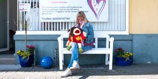 Heike Becker, eine Frau mittleren Alters, sitzt mit einer Handpuppe auf einer Bank vor einem Schild mit der Aufschrift "Seniorenbetreuung mit Herz".