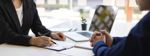 Zwei Personen sitzen an einem Tisch, auf dem ein Laptop steht, und gehen Dokumente durch.