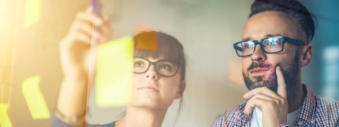 Frau und Mann stehen vor einem Glasboard, auf dem bunte Post-its kleben