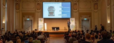 Großer Saal mit Publikum. Ein Mann hält einen Vortrag vor einem großen Bildschirm, der an der Wand hängt.